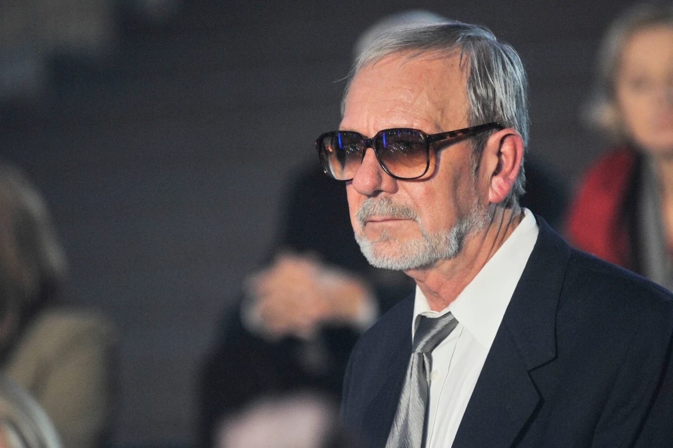 Jerzy Kamas Jerzy Kamas w garniturze i okularach przeciwsłonecznych siedzi na widowni podczas wydarzenia.