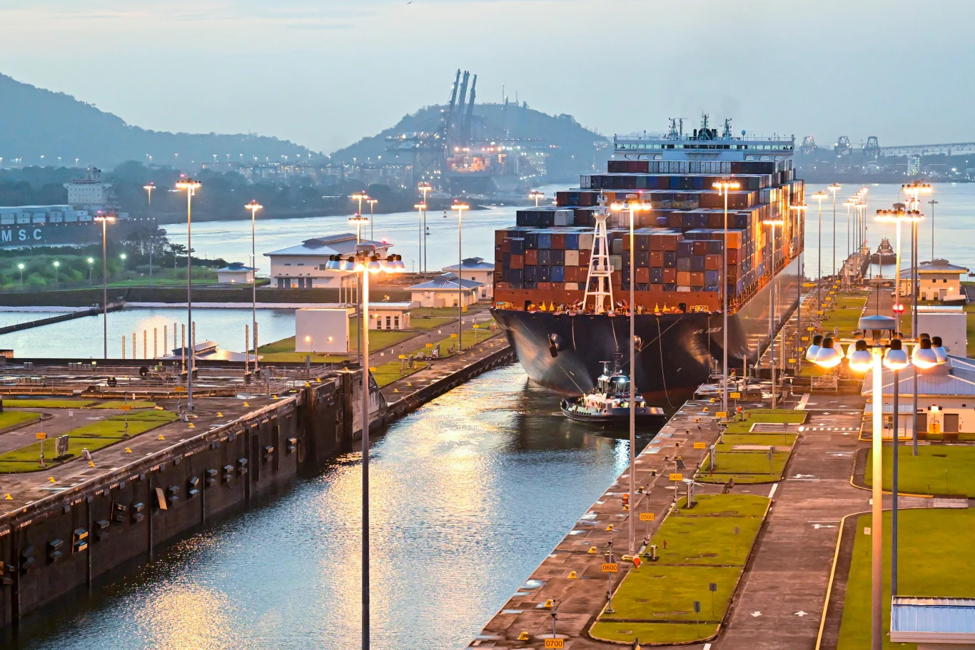 Ceny za prawo do przepłynięcia Kanału Panamskiego osiągnęły rekordowy poziom Śluza kanału. Przepływa przez nią kontenerowiec. Tłem nabrzeże i port. Całość oświetlona przez latarnie.