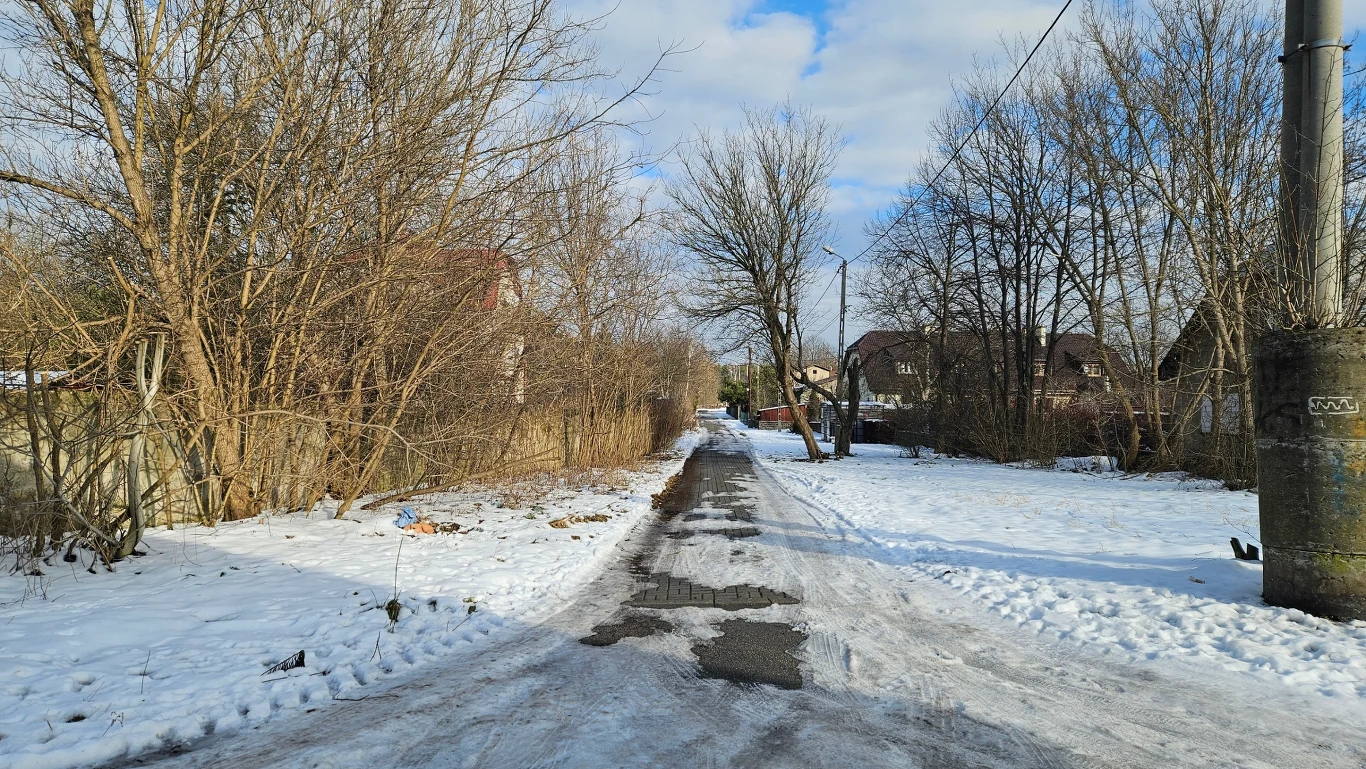 Wąska, zaśnieżona droga wiejska w zimowym krajobrazie, otoczona drzewami bez liści. W tle stoją domy jednorodzinne, a niebo jest częściowo pochmurne. Słup energetyczny po prawej stronie drogi dodaje do kompozycji industrialny akcent.