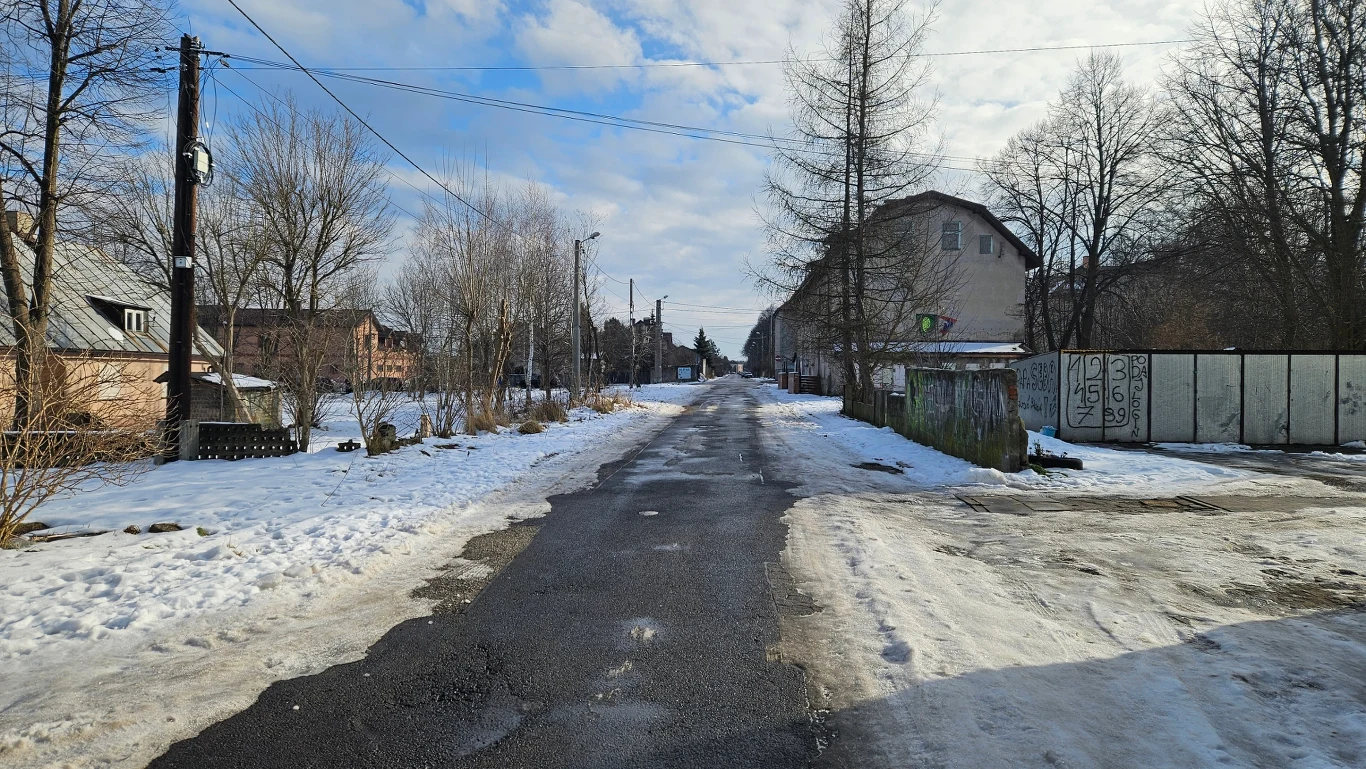Zimowa ulica w małym mieście, otoczona domami i drzewami, ze śniegiem pokrywającym chodniki. W tle widoczna droga asfaltowa ciągnąca się między rzędem budynków po obu stronach i niebo pokryte lekkimi chmurami.