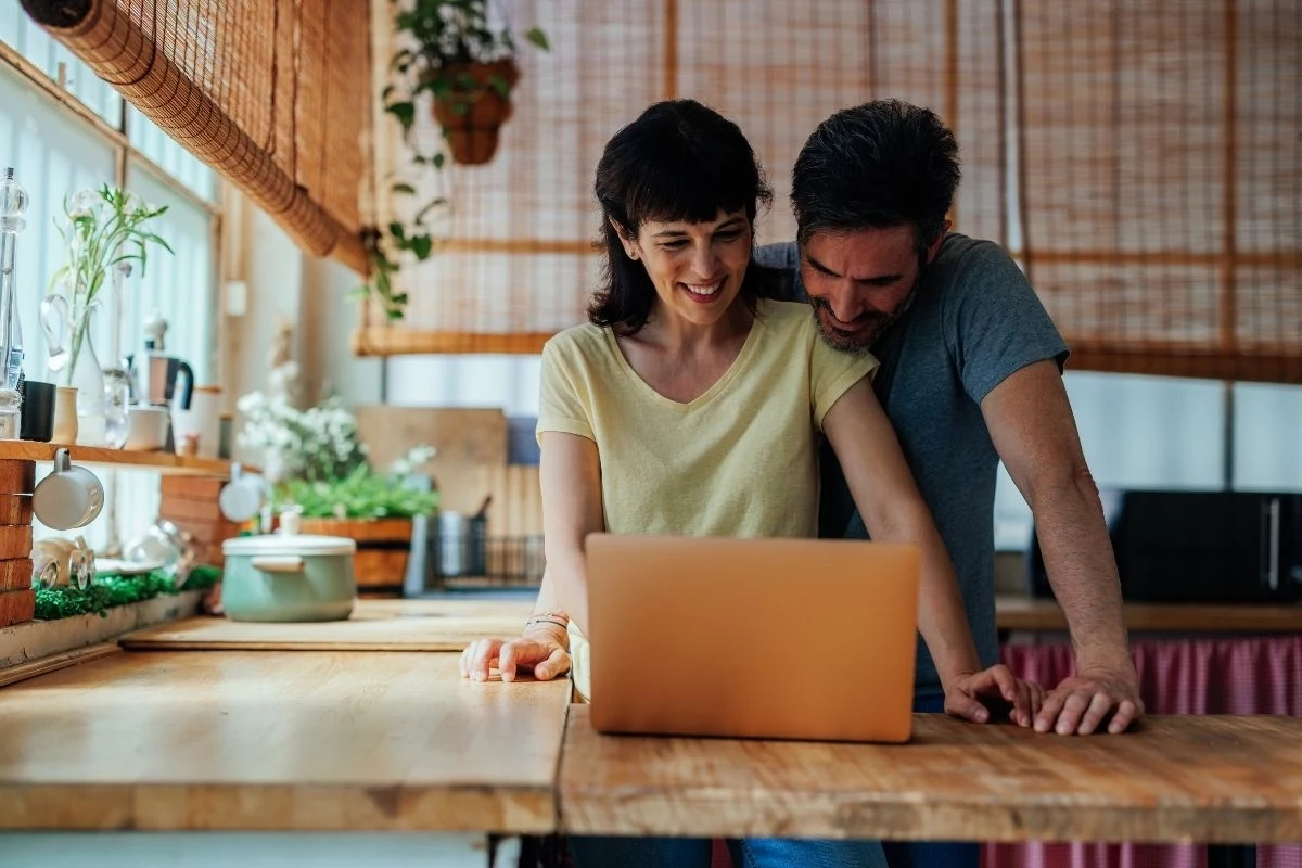 Para stoi blisko siebie w kuchni, z uśmiechem spoglądając na ekran laptopa na blacie. Wnętrze jest jasne i przytulne dzięki naturalnemu światłu wpadającemu przez okno oraz zielonym roślinom w tle.