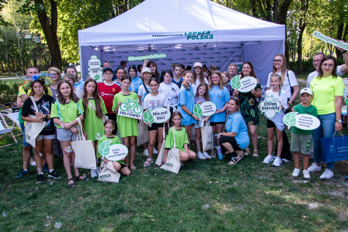 Grupa młodych ludzi przed namiotem z logiem "Lepsza Polska", trzymających tabliczki z napisami promującymi ekologię. Uczestnicy ubrani są w jasne kolory, a otoczenie jest zielone, co nadaje scenie przyjemny, naturalny charakter.