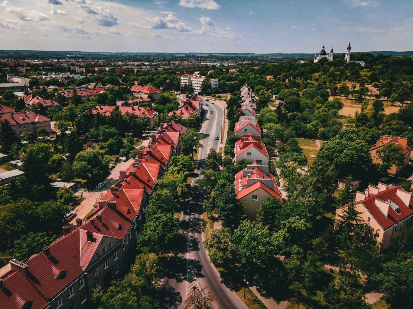 Panoramiczne ujęcie bloku mieszkalnego z czerwonymi dachami i otaczającą zielenią. Na horyzoncie widoczny jest zarys kościoła i zabudowa miejska. Droga biegnie środkiem zdjęcia, prowadząc w głąb miasteczka. Chmury zdobią błękitne niebo.