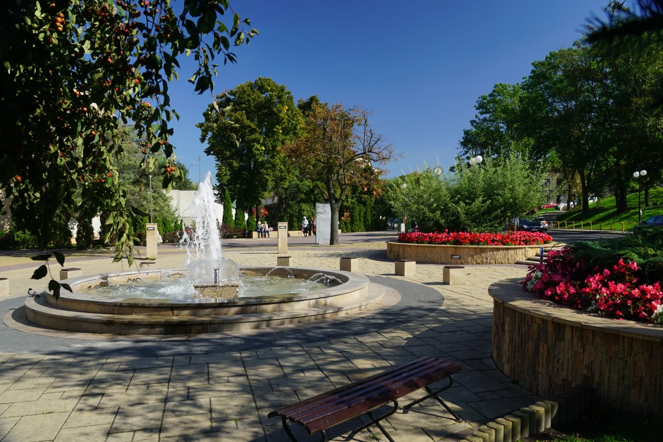 Fontanna na placu otoczona zielenią i kolorowymi kwiatami. Ławeczki i alejki dookoła zachęcają do odpoczynku. Promienie słońca oświetlają przestrzeń, podkreślając uroki spokojnego miejsca w parku.