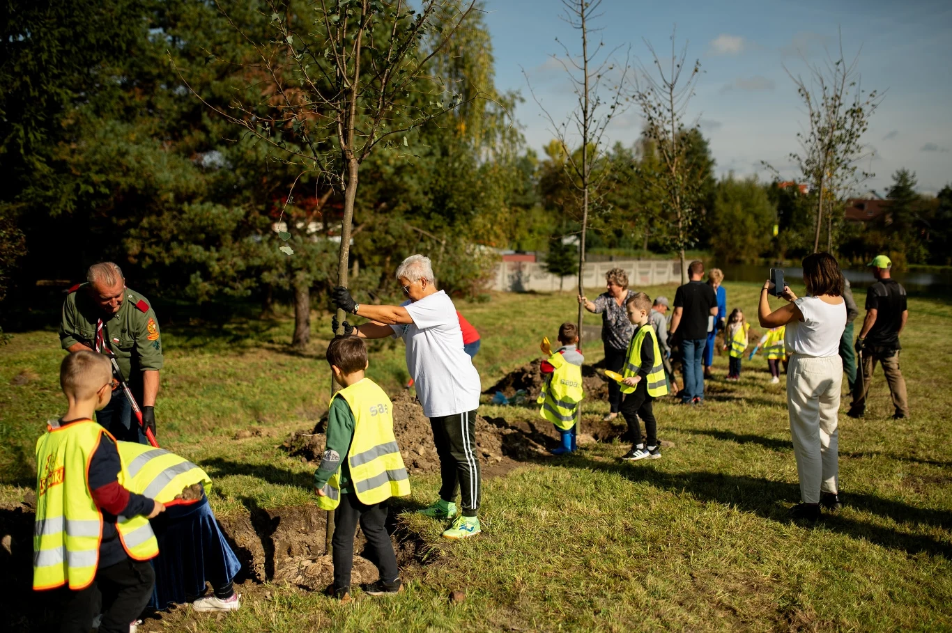 Czyste Zielone Miasta fot. UM Chrzanów Grupa dorosłych i dzieci w odblaskowych kamizelkach sadzi drzewa w parku. W tle widać drzewa i trawnik. Ludzie są zaangażowani w akcję sadzenia, a część z nich robi zdjęcia.