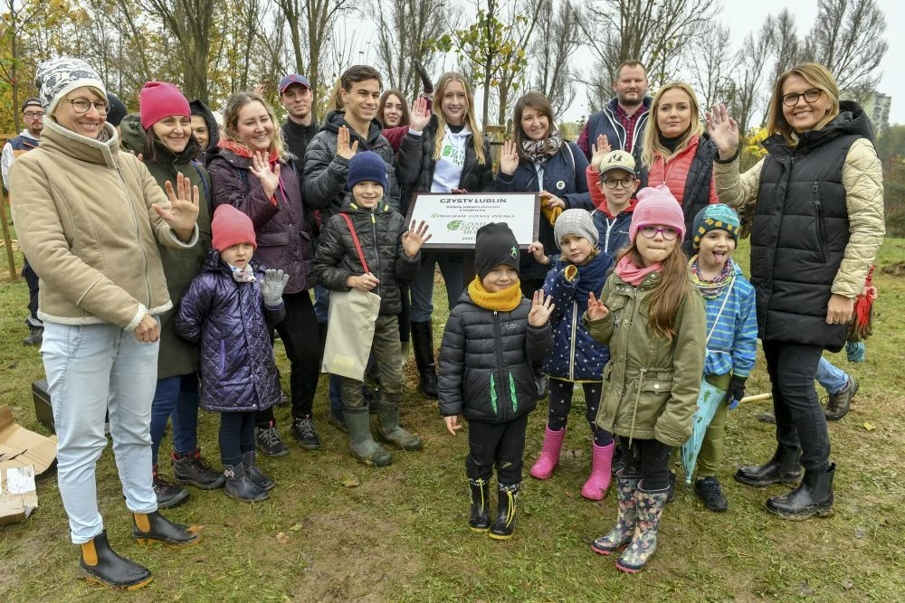Czyste Zielone Miasta fot. Telewizja Polsat Uśmiechnięta grupa ludzi, składająca się z dzieci i dorosłych, stoi na trawie w parku. W centrum trzymają certyfikat. Wszyscy są ciepło ubrani w zimowe kurtki i czapki, sugerując chłodną pogodę.