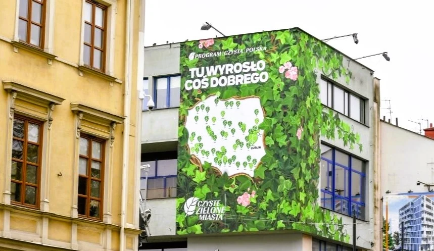 Czyste Zielone Miasta fot. Telewizja Polsat Kolorowy mural na ścianie miejskiego budynku przedstawiający mapę Polski, której kontury wypełniają zielone liście i różowe kwiaty. Obok znajdują się napisy promujące ekologiczny program na rzecz czystości i zieleni w miastach.