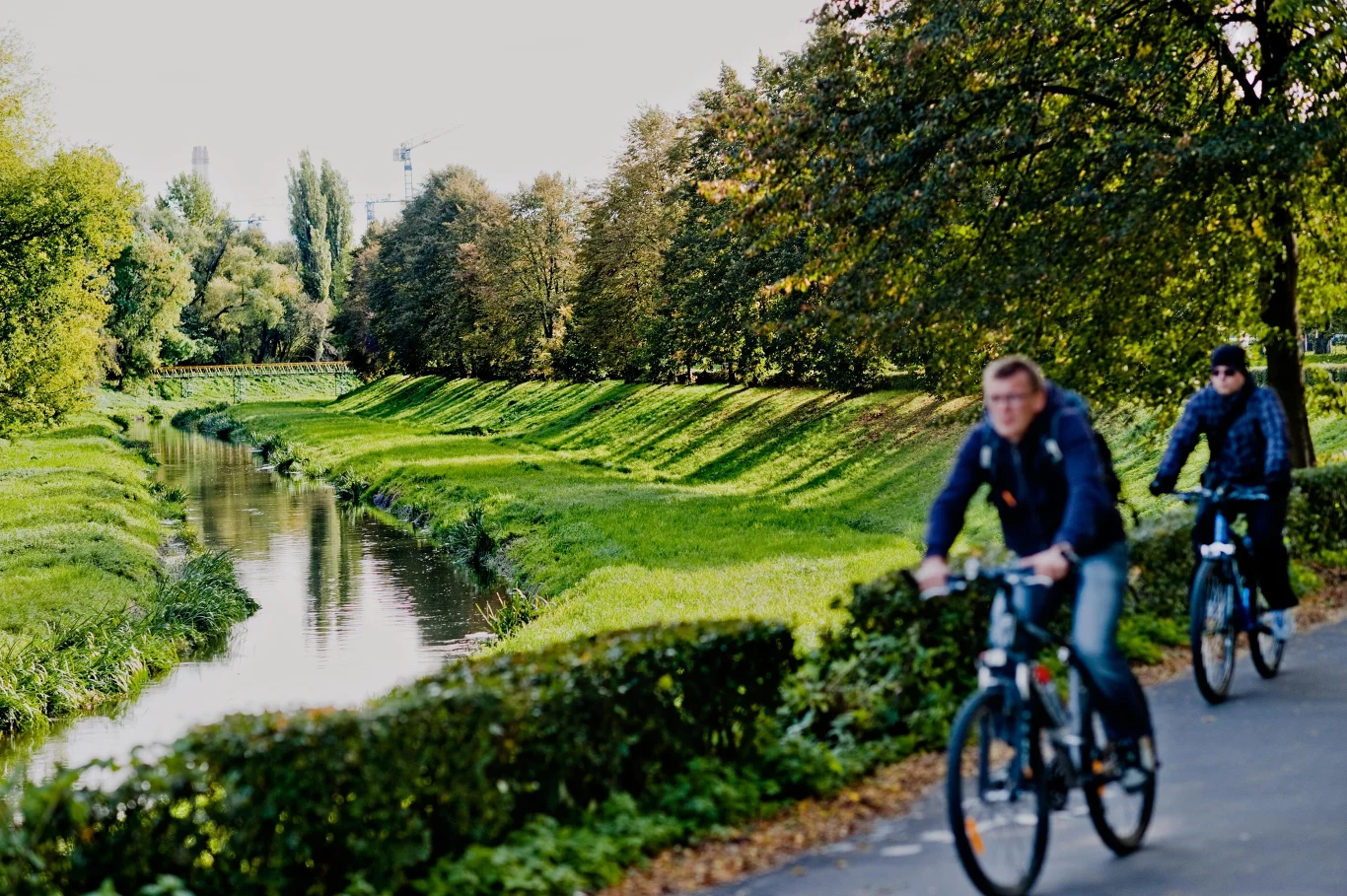 Czyste Zielone Miasta fot. UM Lublin Dwaj rowerzyści jadą ścieżką wzdłuż zielonego kanału, otoczonego drzewami. Znajdują się w ruchu, w pogodnym i naturalnym otoczeniu.