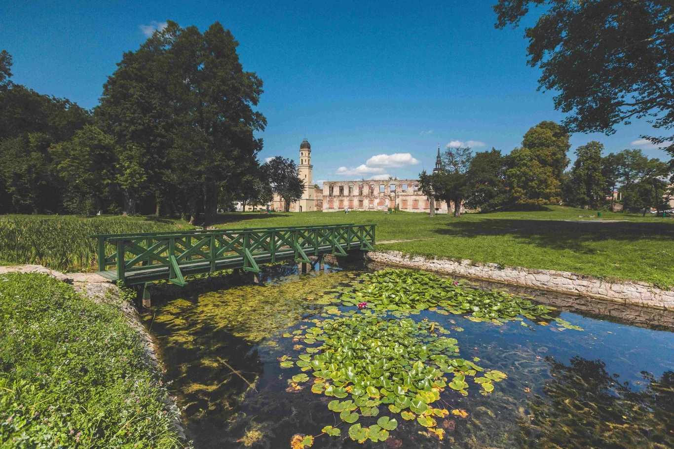 Malowniczy widok pałacu w tle, otoczonego przez bujny park z małym mostkiem nad stawem pełnym lilii wodnych, otoczony wysokimi drzewami.