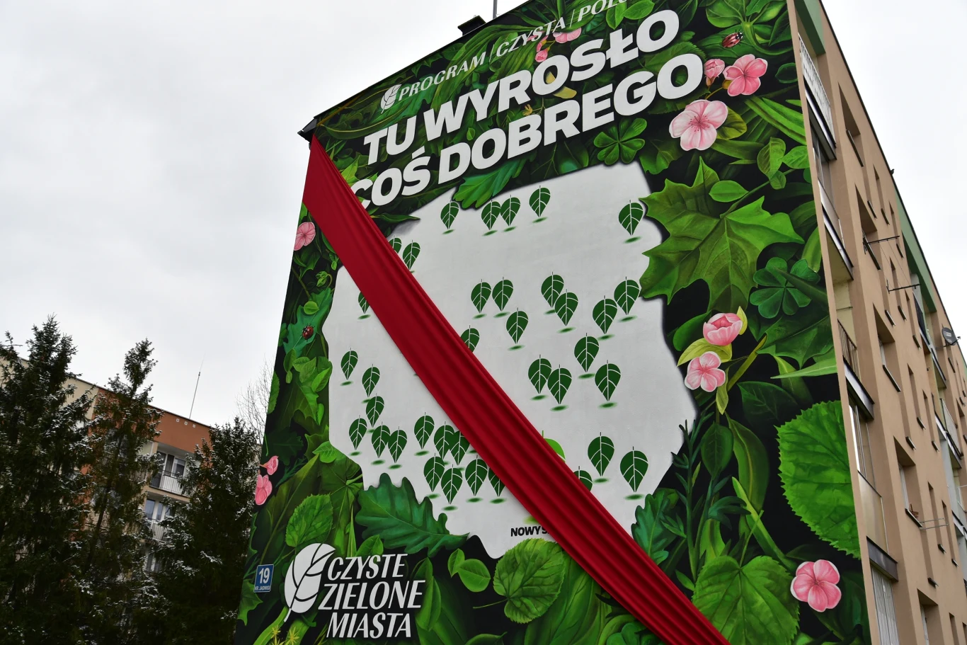Mural na ścianie budynku z napisem 'TU WYROSŁO COŚ DOBREGO', otoczony motywami roślinnymi z mapą Polski w tle. Po lewej stronie widoczne są drzewa. Na dole znajduje się logotyp 'Czyste Zielone Miasta'. U dołu budynku zaczyna się czerwona wstęga.