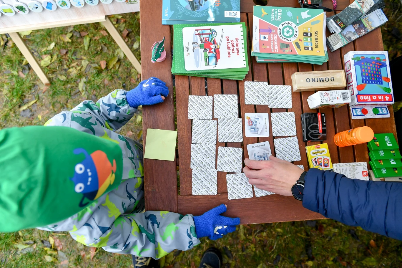 Dziecko w zielonej czapce i rękawiczkach gra w grę memory przy stole z różnymi książkami, kartami i zabawkami. Ręka dorosłego doradza w grze.