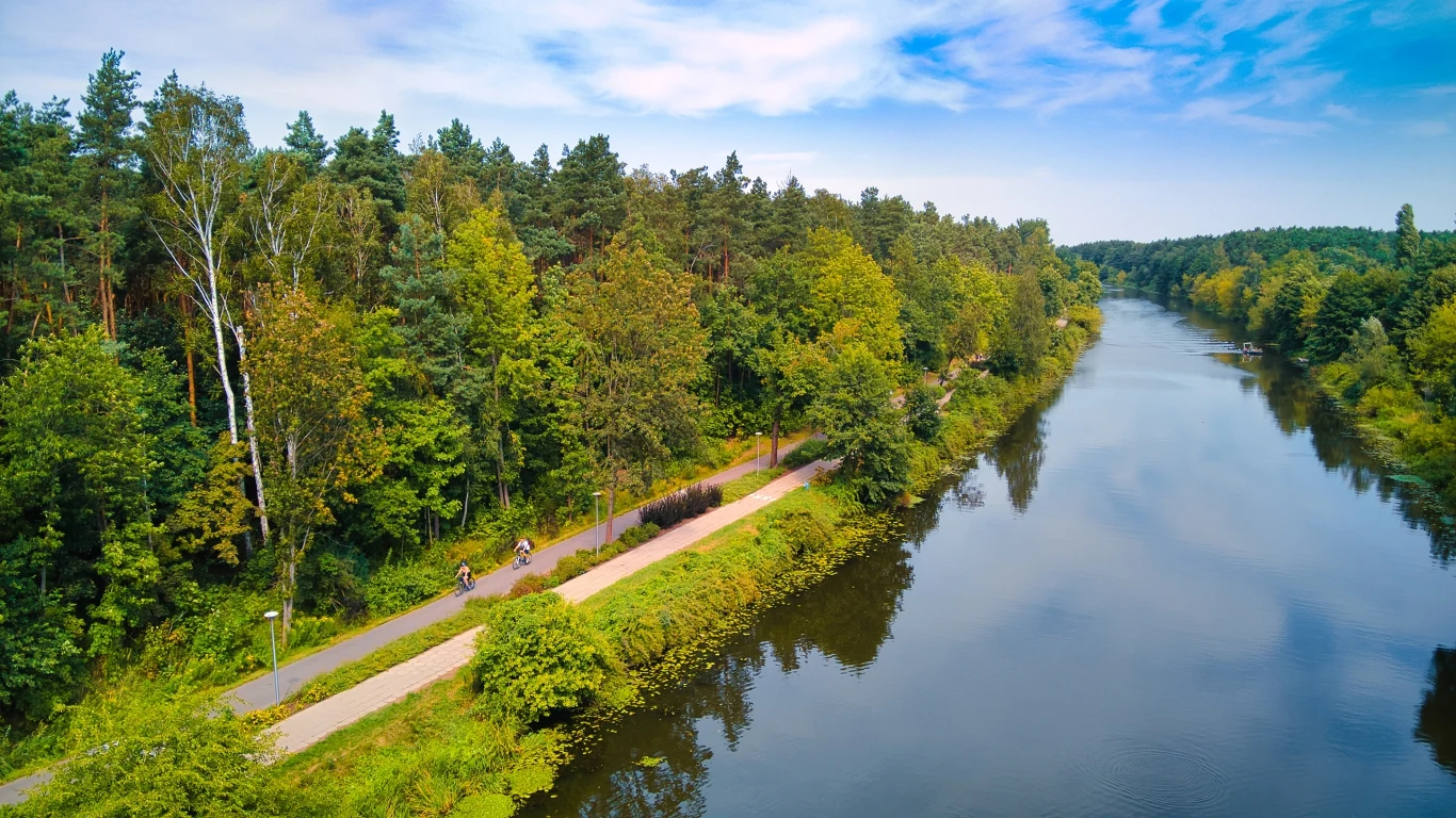 Widok z lotu ptaka na malowniczy zakątek przyrody z rzeką biegnącą wzdłuż lasu, ścieżce rowerowej prowadzącej przez zalesiony teren oraz kilkoma rowerzystami cieszącymi się aktywnością na świeżym powietrzu.