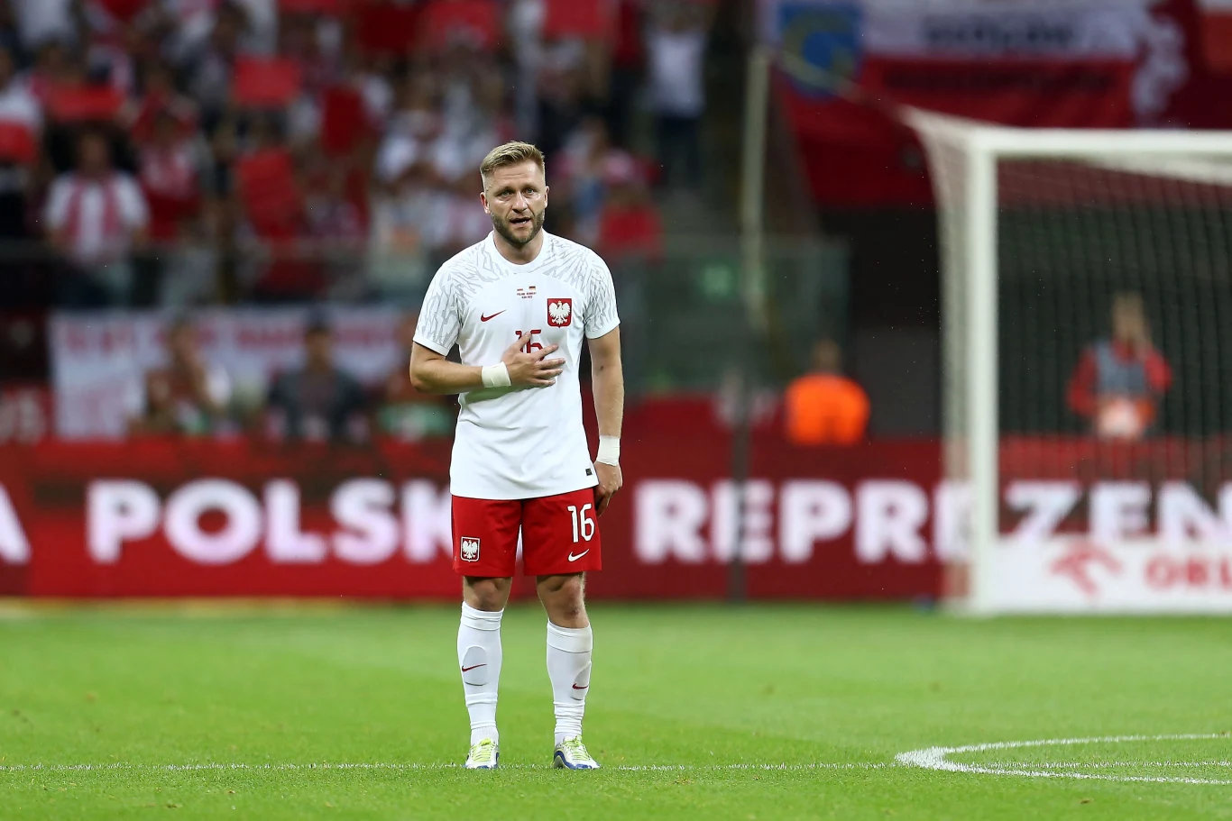 Jakub Błaszczykowski Jakub Błaszczykowski w stroju reprezentacji Polski stoi na murawie, w tle kibice z flagami narodowymi.
