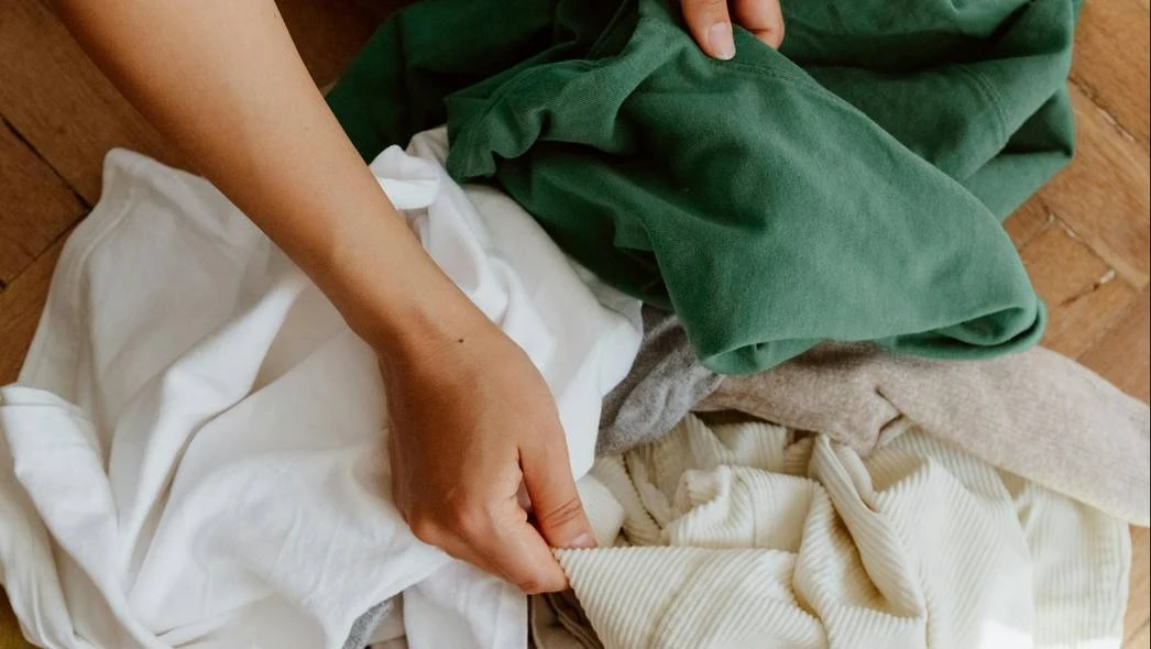 Ręce przeglądające stos ubrań w różnych kolorach, skupiając się na zielonej koszuli. Ubrania leżą na drewnianej podłodze, a ręce są w ruchu, jakby sortowały rzeczy.