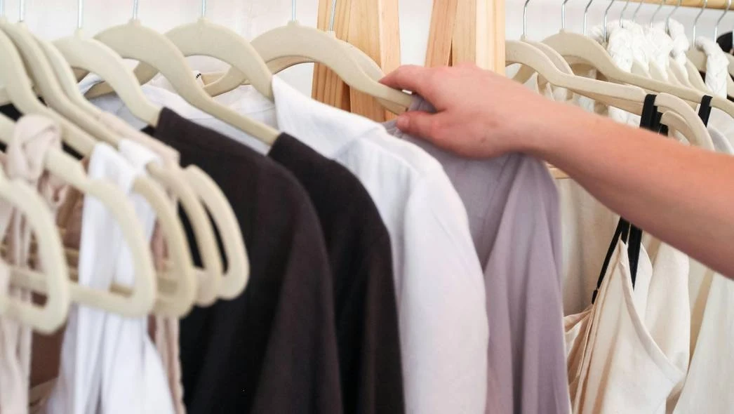 Ręka przeglądająca różnorodne ubrania wiszące na wieszakach, w tym białe, czarne i szare bluzki umieszczone na beżowych wieszakach.
