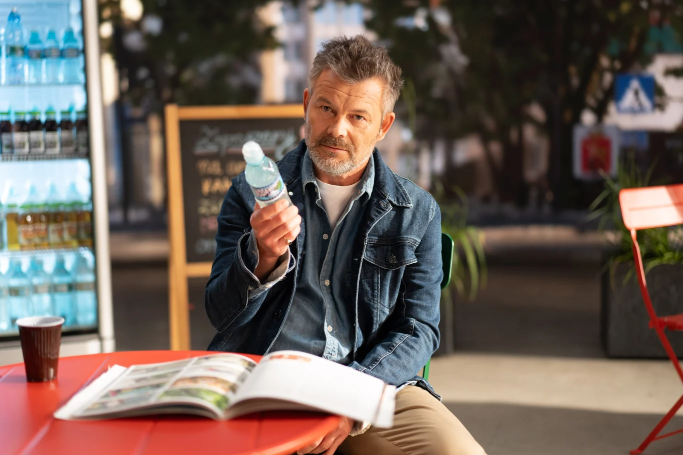 Jacek Braciak w spocie Stowarzyszenia - Fot. ATM Virtual Mężczyzna w dżinsowej kurtce siedzi przy czerwonym stoliku na zewnątrz i trzyma butelkę wody mineralnej. Na stole znajduje się otwarta książka i papierowy kubek z kawą. W tle lodówka z napojami i tablica ogłoszeń.