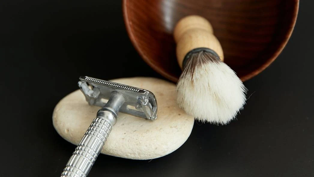 Tradycyjna maszynka do golenia leży na jasnym kamieniu, obok pędzel do golenia z drewnianą rączką oraz brązowa miseczka. Akcesoria znajdują się na czarnym tle.