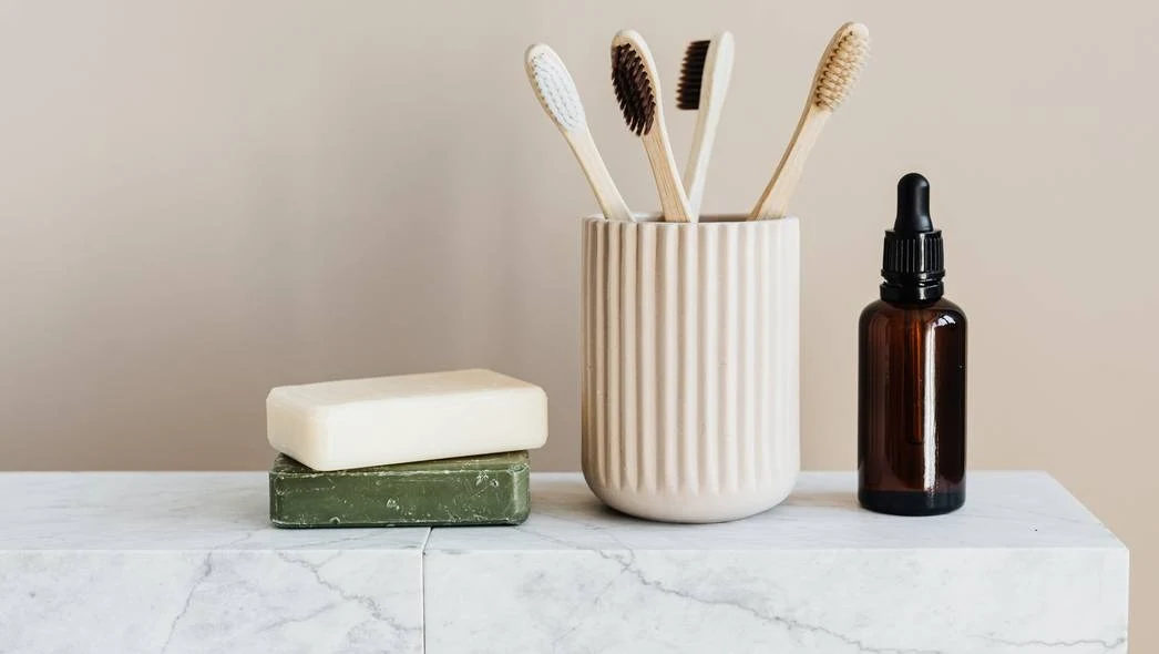 Zestaw przyborów łazienkowych na marmurowym blacie, obejmujący bambusowe szczoteczki do zębów w ceramicznym kubku, dwa mydła w kostce jedno na drugim oraz brązowa buteleczka z pipetą wypełniona płynem.