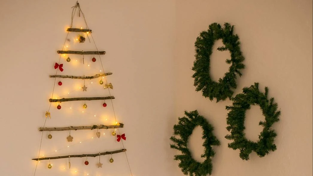 Fot. pexels.com/ Юлия Здобнова/ Dekoracje świąteczne na ścianie, po lewej stronie drzewko z gałęzi i lampek, ozdobione bombkami i kokardkami, po prawej trzy wieńce z zielonych gałązek.