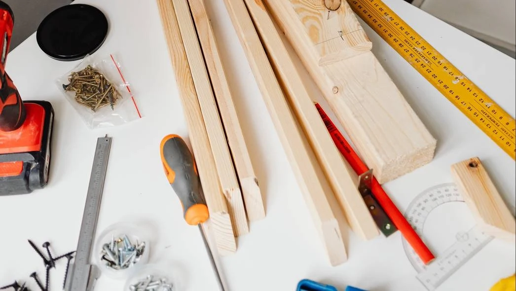 Narzędzia stolarskie i materiały, takie jak drewniane belki, wkrętarka, miara i śruby ułożone na stole.