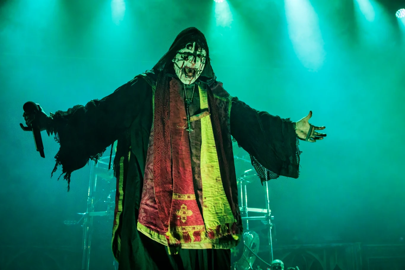 Attila Csihar (Mayhem) w akcji Artysta stojący na scenie w teatralnym kostiumie i makijażu, trzyma mikrofon. Wszystko w zielonym oświetleniu, wewnątrz widoczne elementy bębnów perkusyjnych.