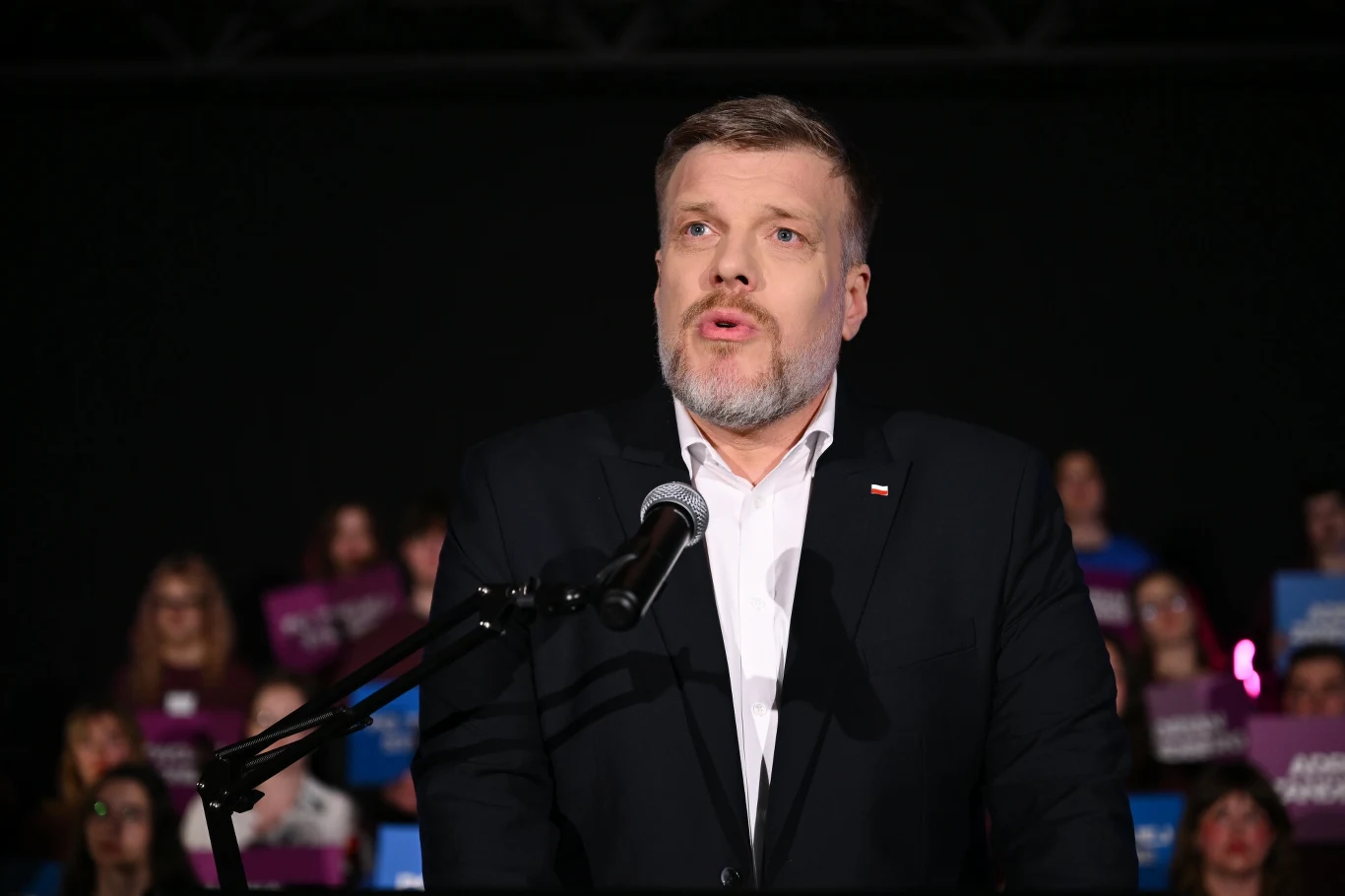 Adrian Zandberg, kandydat Razem w wyborach prezydenckich Mężczyzna w garniturze przemawia do mikrofonu na tle osób z plakatami, prawdopodobnie podczas wydarzenia publicznego.