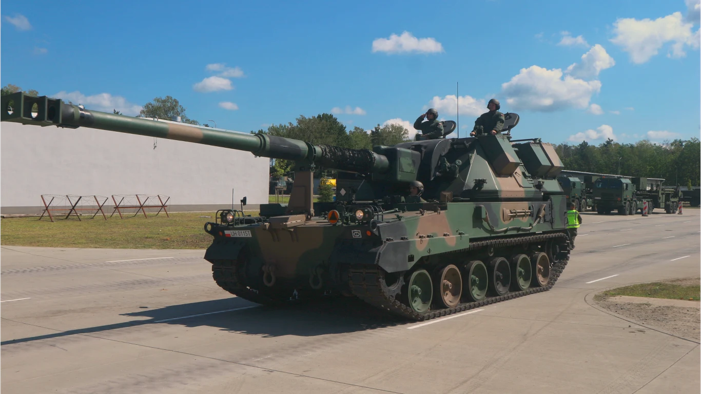 Pojazd wojskowy, prawdopodobnie armata samobieżna, porusza się po asfaltowej drodze. Na jego powierzchni widoczny jest wzór kamuflażu, a na wieży stoją żołnierze. W tle drzewa i niebo z chmurami, co sugeruje spokojne warunki atmosferyczne.