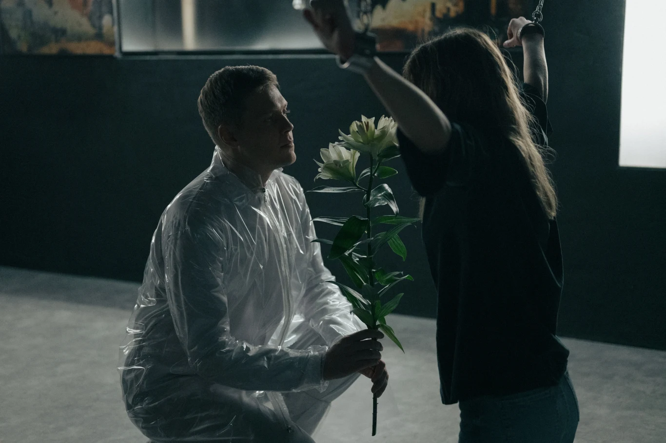 Mężczyzna w białym kombinezonie ochronnym klęczy przed kobietą, trzymając dużą, białą lilię. Kobieta o długich włosach ma ręce podniesione do góry. Scena jest ciemna i tajemnicza, z jasnym światłem oświetlającym postacie.