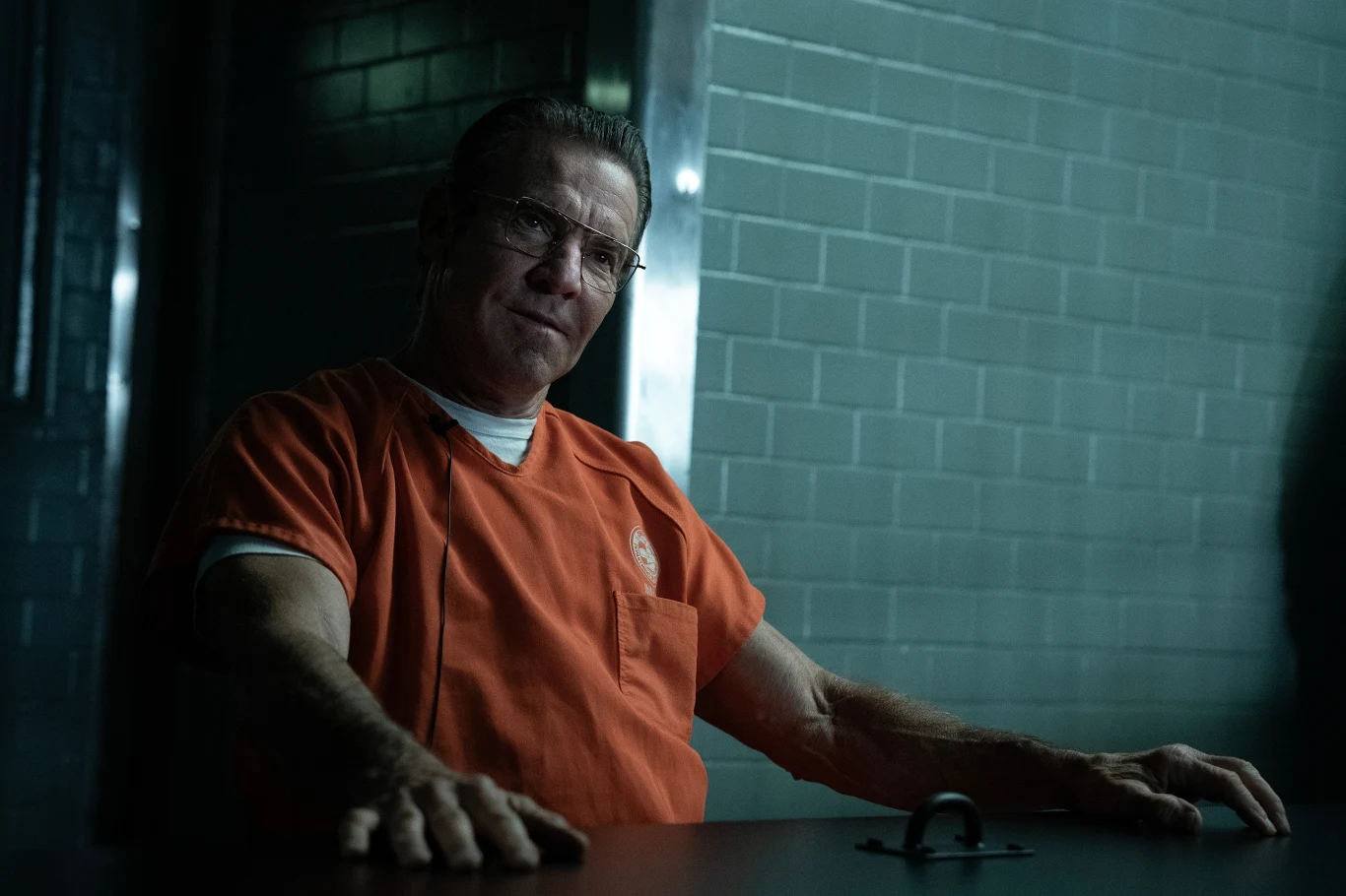 Mężczyzna w pomarańczowym kombinezonie więziennym siedzi przy metalowym stole w ciemnym pomieszczeniu, przypominającym celę przesłuchań. Ściany pokryte są szarymi kafelkami, a oświetlenie tworzy surową atmosferę.