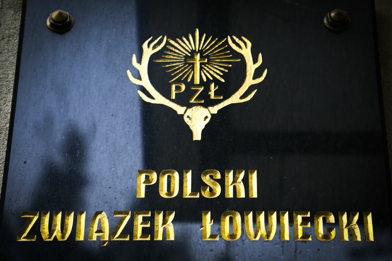 Tablica z logo Polskiego Związku Łowieckiego, złote litery na ciemnym tle, centralnie umieszczony emblemat w kształcie rogów jelenia z krzyżem.