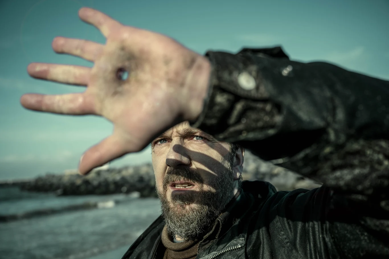 Mężczyzna nad brzegiem morza z uniesioną ręką, na której jest widoczna dziura, co rzuca cień na jego twarz.