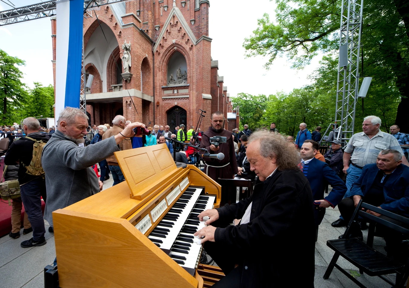 Józef Skrzek prezentuje pierwszy z trzech albumów szykowanych na 2025 r. Mężczyzna grający na organach elektronicznych na tle gotyckiego kościoła, otoczony przez publiczność na świeżym powietrzu. Wiele osób obserwuje występ, a atmosfera jest żywa i pełna zainteresowania.