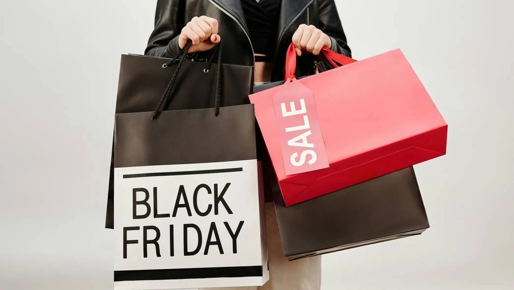 Torby zakupowe w rękach osoby trzymającej czarną torbę z napisem "BLACK FRIDAY" oraz czerwoną z napisem "SALE" na jasnym tle.
