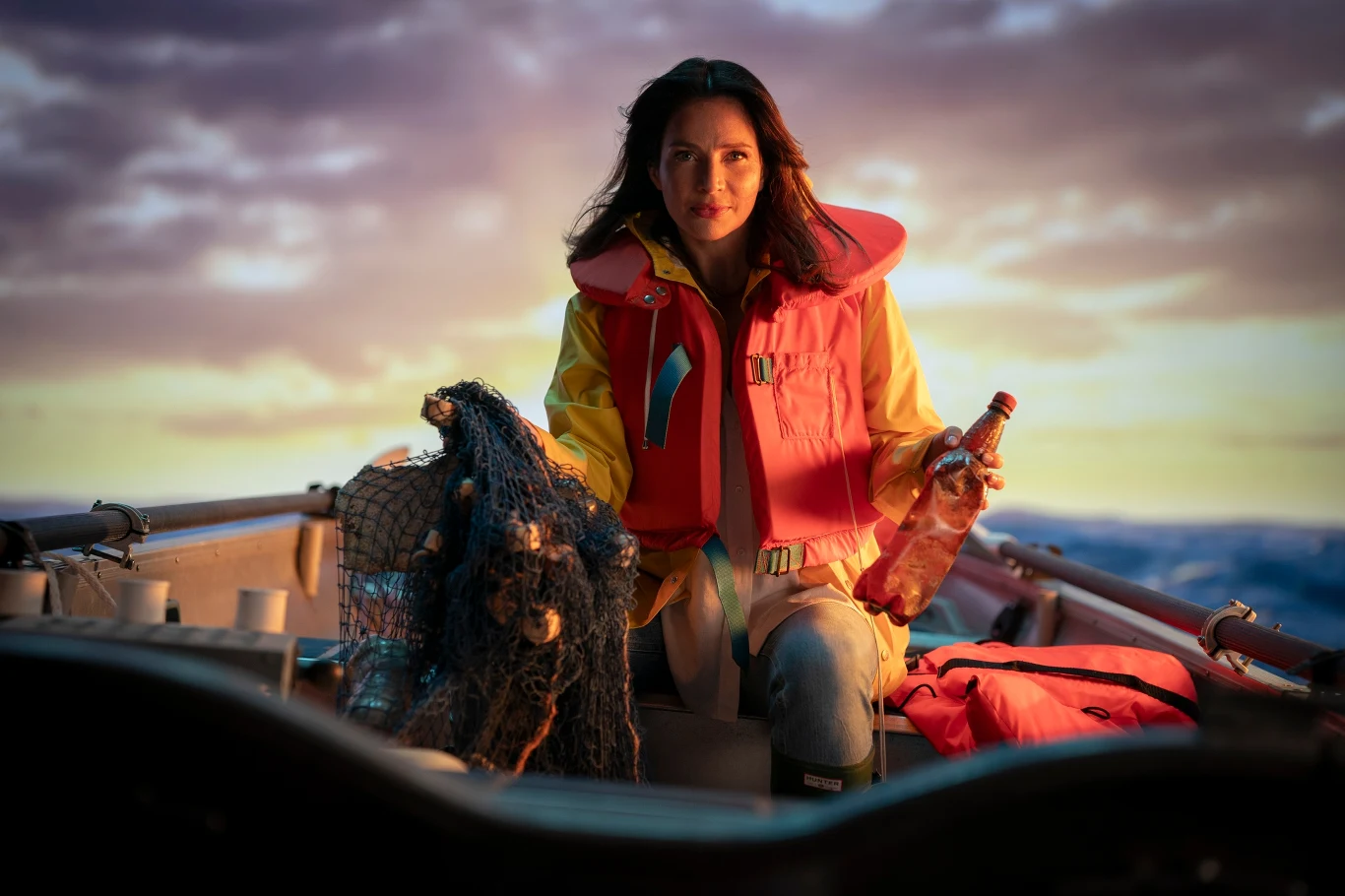 Kobieta w kamizelce ratunkowej siedzi na łodzi, trzymając butelkę w dłoni. Obok niej znajduje się sieć rybacka, sugerująca motyw morskiej wyprawy. Tło stanowią rozległe, wzburzone fale i zachmurzone niebo, co wprowadza dramatyczny nastrój.