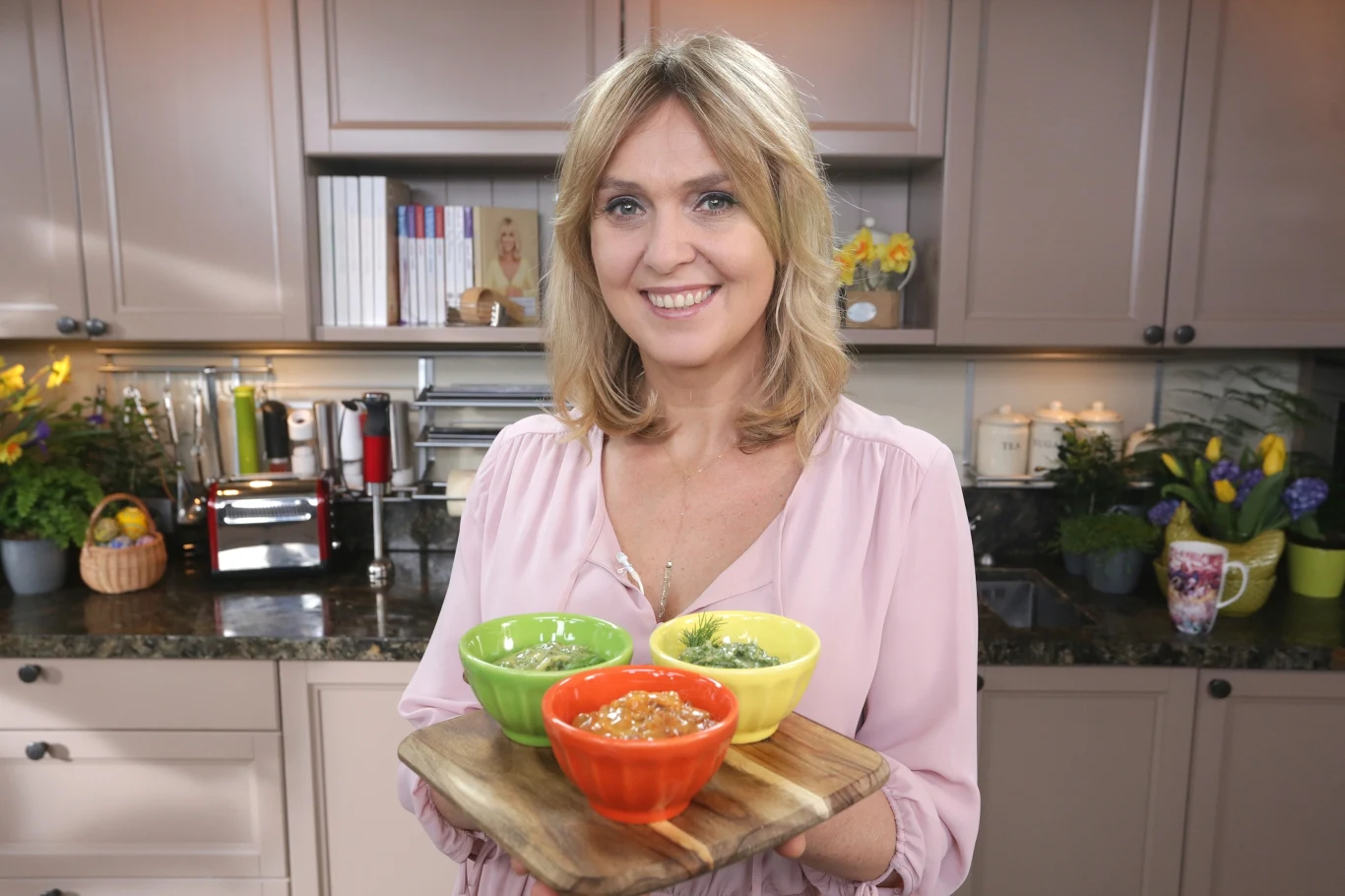 Ewa Wachowicz poleca dodatki do wielkanocnych jajek Kobieta w kuchni trzyma drewnianą deskę z trzema kolorowymi miseczkami, w których znajdują się różne potrawy. Uśmiechnięta, nosi różową bluzkę, w tle widać kuchenne szafki oraz dekoracje.