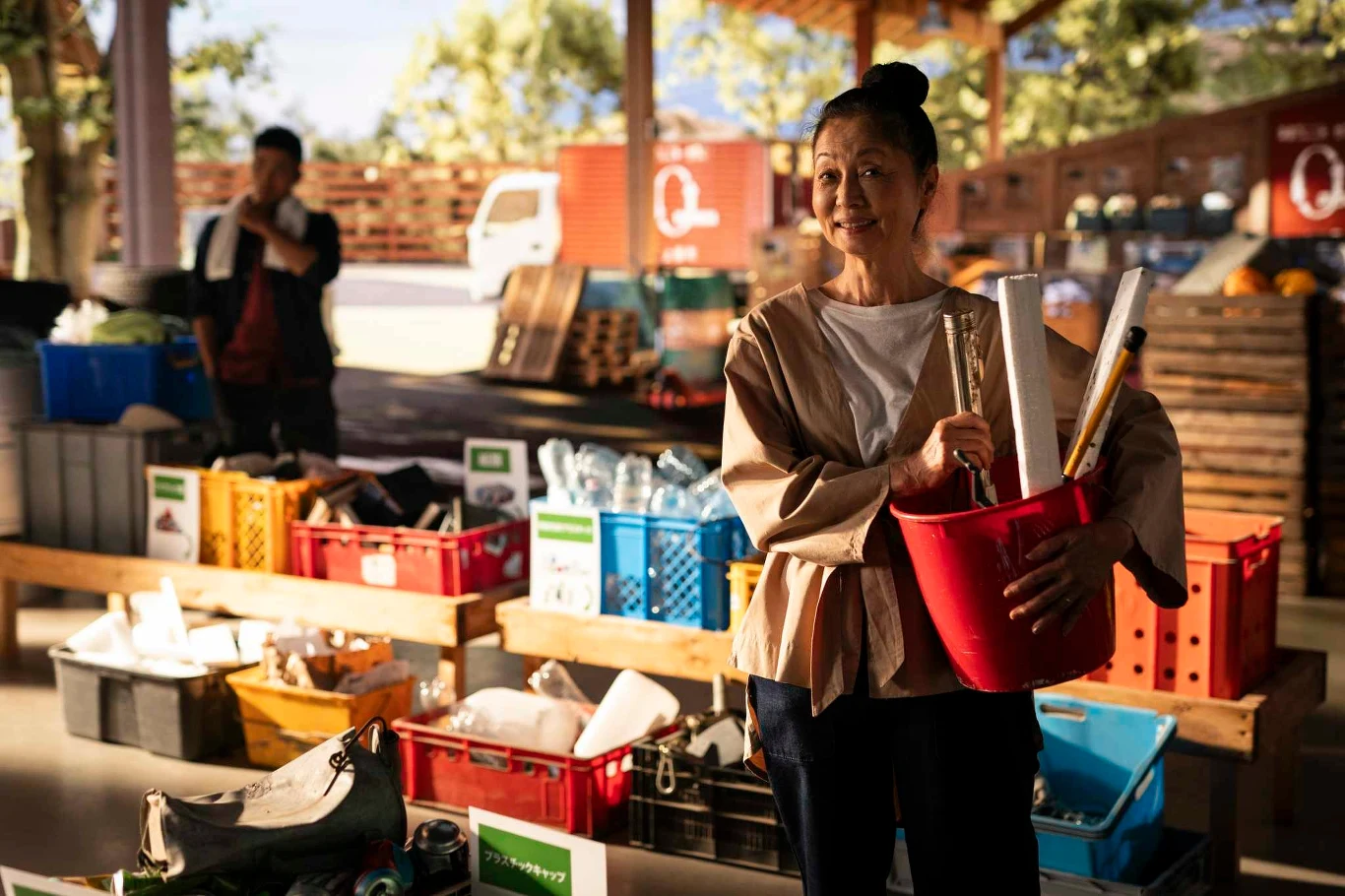 Kobieta trzymająca czerwone wiadro z materiałami, stojąca przed stołem pełnym pojemników wypełnionych różnymi przedmiotami. W tle widać drugą osobę oraz zaparkowany samochód dostawczy.