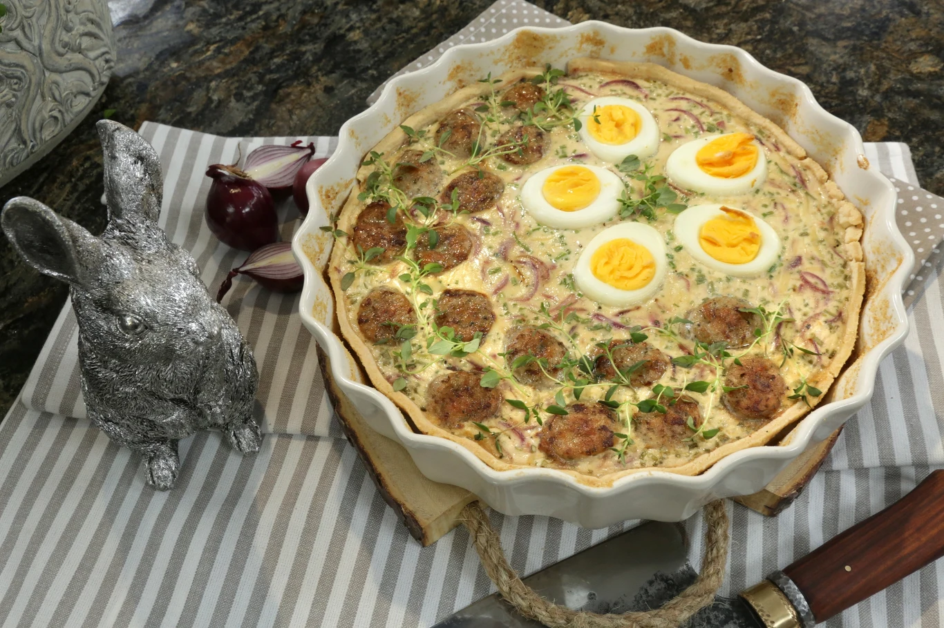 Tarta jak mazurek Ceramiczna forma z wykwintną zapiekanką, ozdobioną plasterkami jajek i świeżymi ziołami, na tle eleganckiej tkaniny w paski. Obok metalowa figurka królika i cebula czerwona.