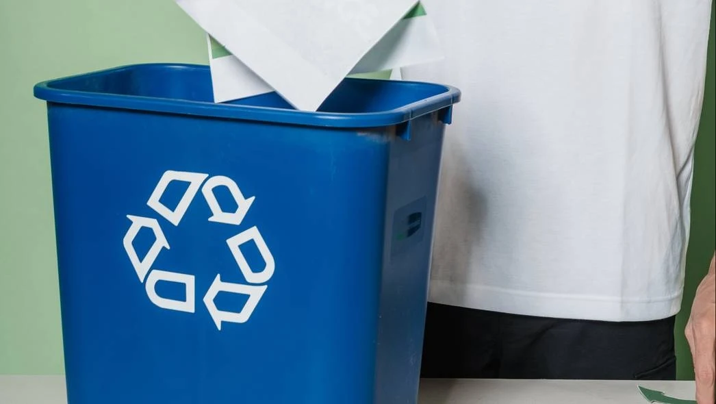 Niebieski pojemnik na śmieci z symbolem recyklingu, do którego ktoś wkłada papierowy dokument. Obok pojemnika widoczna jest osoba w białej koszulce.
