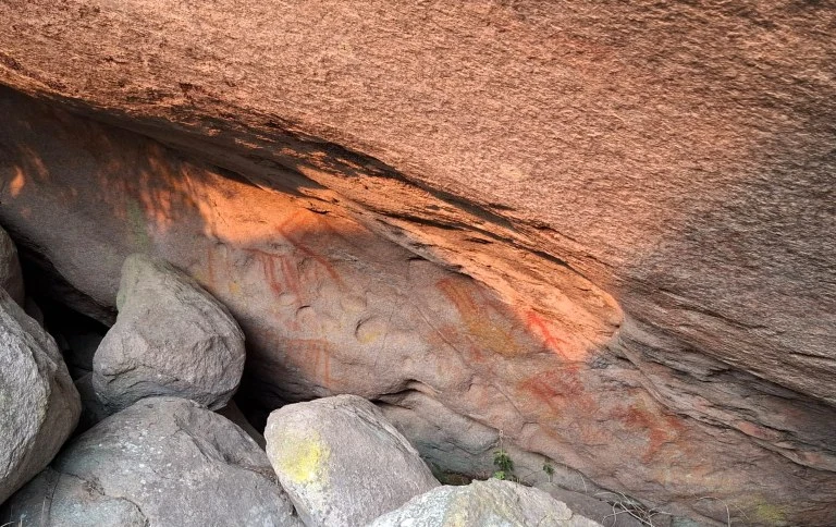 Jedne z malowideł naskalnych Parku Narodowego Itatiaia Skała z prehistorycznymi malowidłami naskalnymi w odcieniach pomarańczowych i czerwonych, otoczona większymi kamieniami.