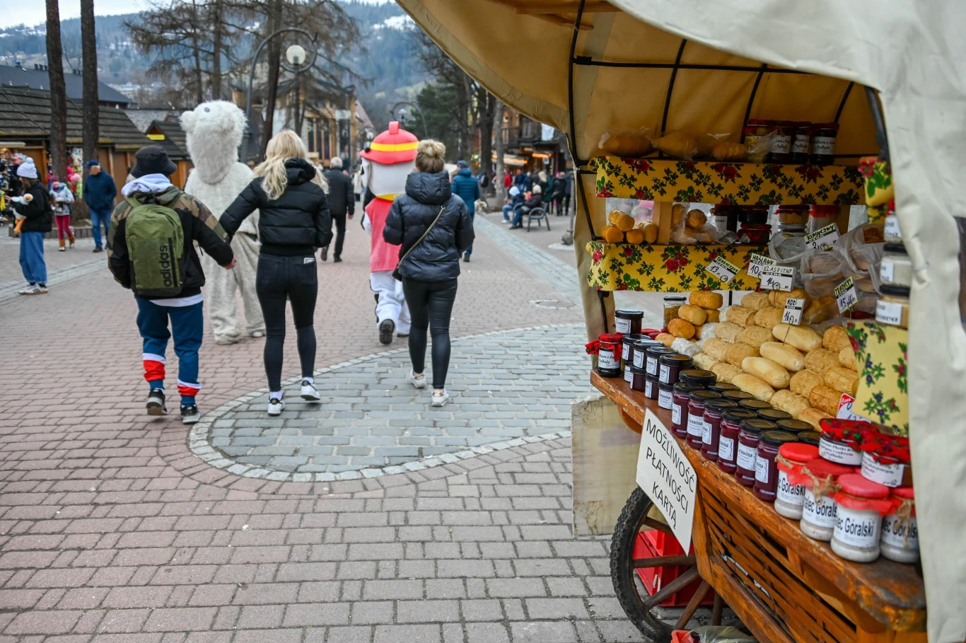 Turyści zjadą raczej na majówkę Budka z regionalnymi produktami, takimi jak oscypki i słoiki z konfiturami, znajduje się na deptaku w górskim kurorcie. Przechodnie, w tym osoba przebrana za białego misia, spacerują po brukowanej ulicy.
