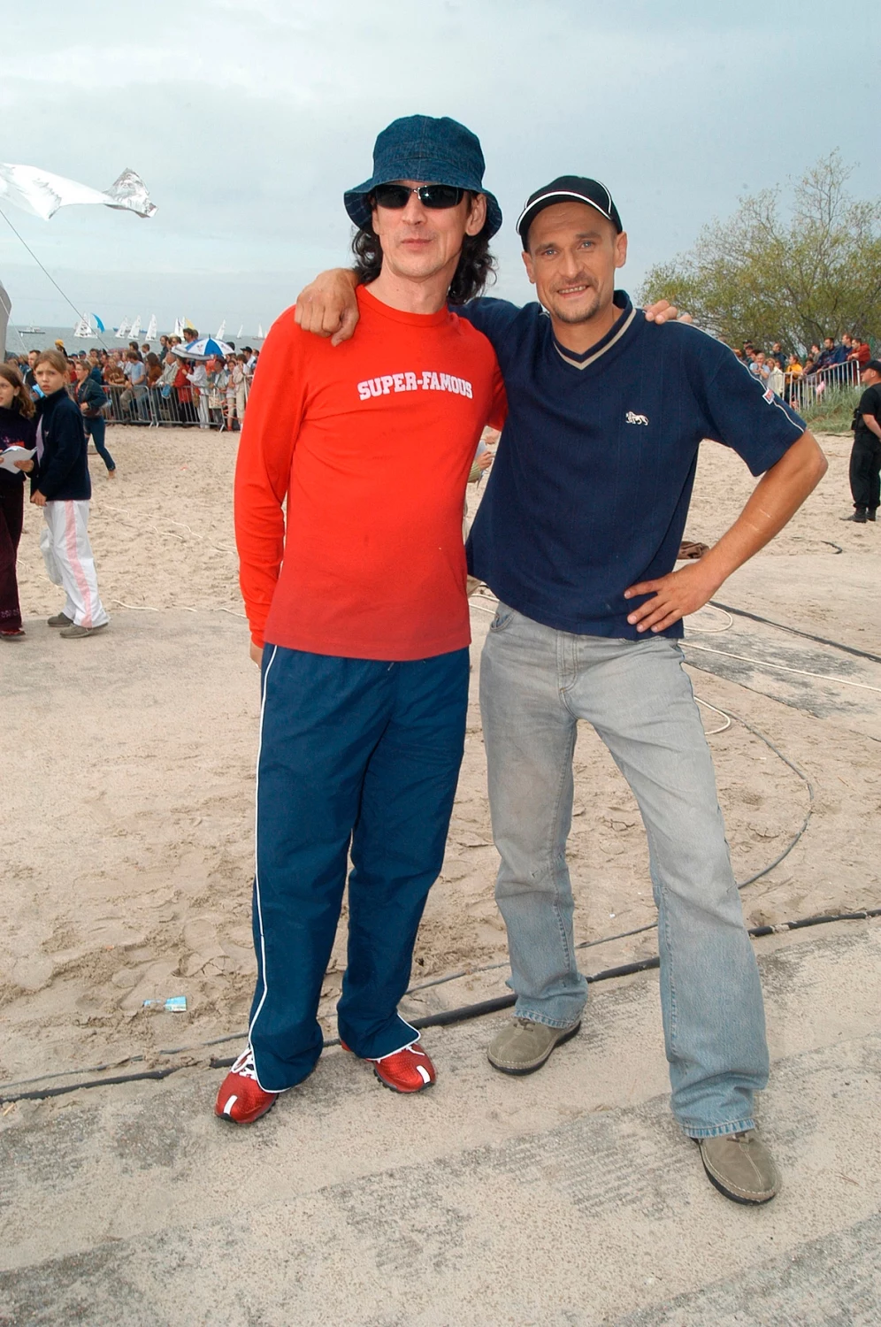 Jan Borysewicz i Paweł Kukiz w 2003 r. Dwóch mężczyzn pozuje na piaszczystej plaży, obaj uśmiechnięci i swobodnie ubrani. Pierwszy z nich ma na sobie czerwoną bluzę i ciemnoniebieskie spodnie, drugi nosi ciemny t-shirt i jasne jeansy. W tle widać grupę ludzi oraz plażowe drzewa.