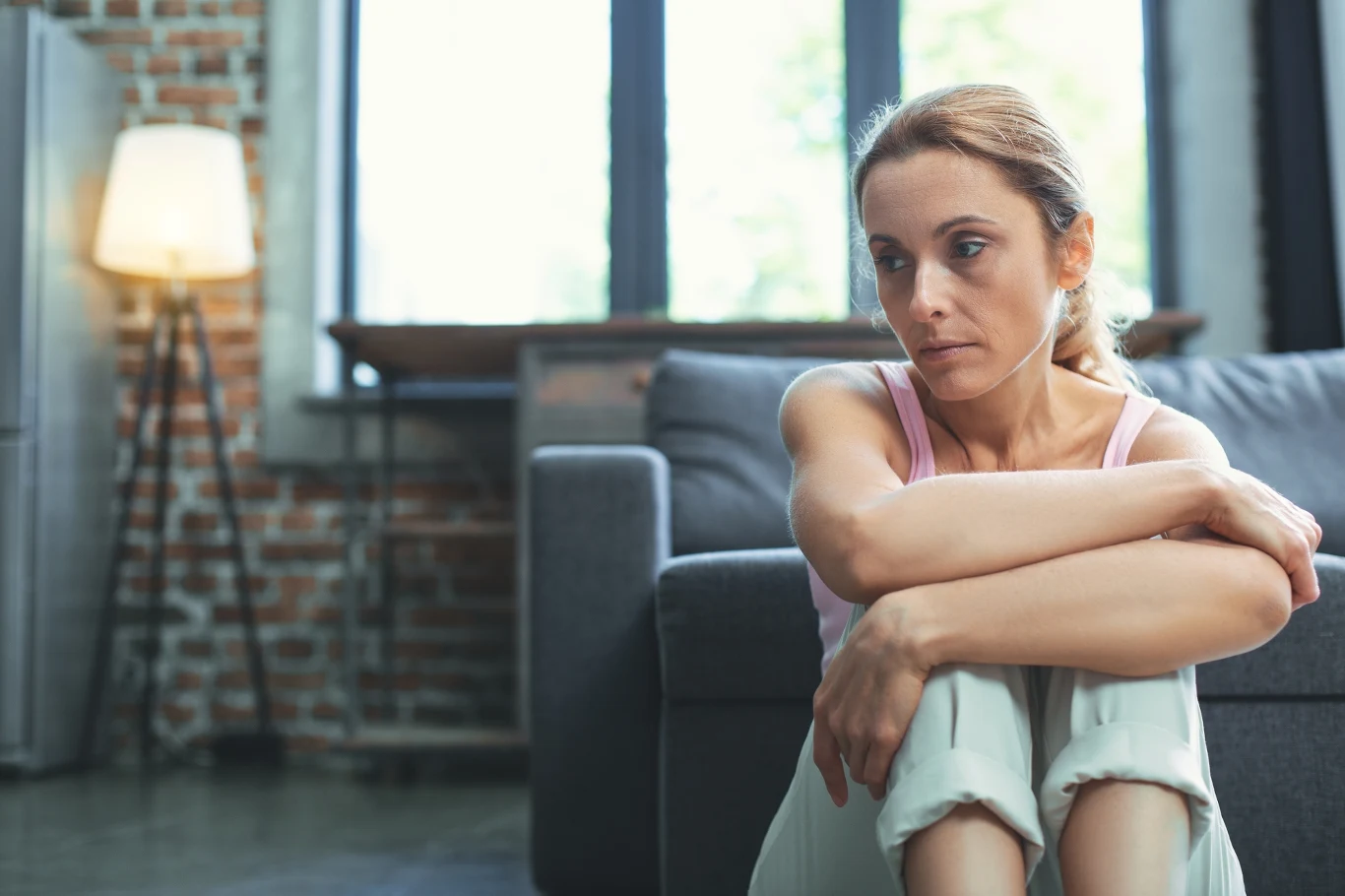 Słowa "kocham cię" mogą budzić różne emocje Kobieta siedząca na podłodze w salonie, wygląda na zamyśloną. Ma jasne włosy związane w kucyk i nosi jasny top oraz spodnie. W tle widać szary narożnik i lampę stojącą, a za oknem wpada naturalne światło.