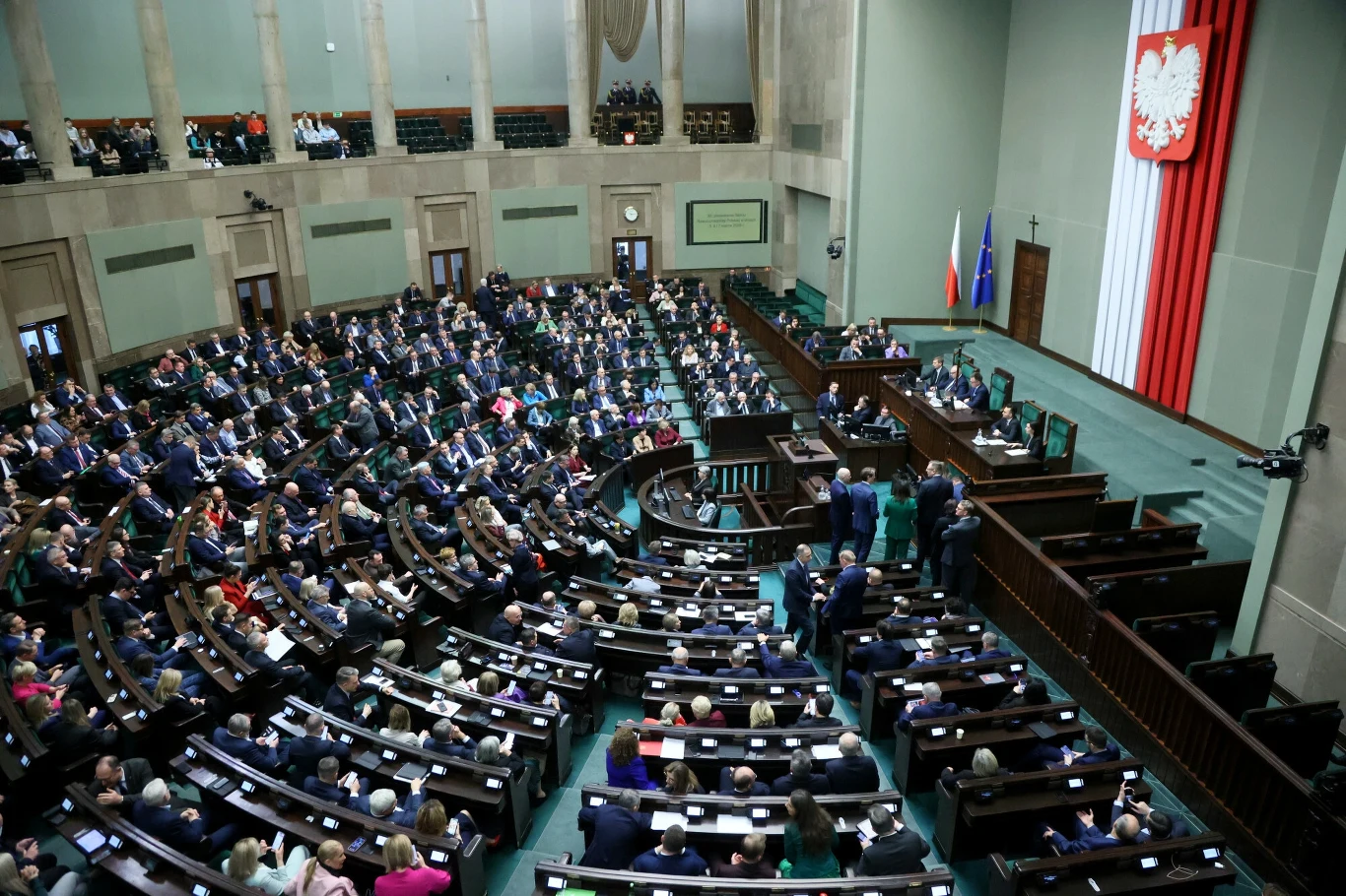 Sala obrad Sejmu. Kto dostałby się do nowego parlamentu? Jest nowy sondaż Sesja parlamentarzystów w sali posiedzeń, z widokiem na mównicę oraz liczne miejsca siedzące wypełnione osobami ubranymi formalnie. Na ścianie widoczna flaga Polski i godło, a z boku stoją flagi narodowe.