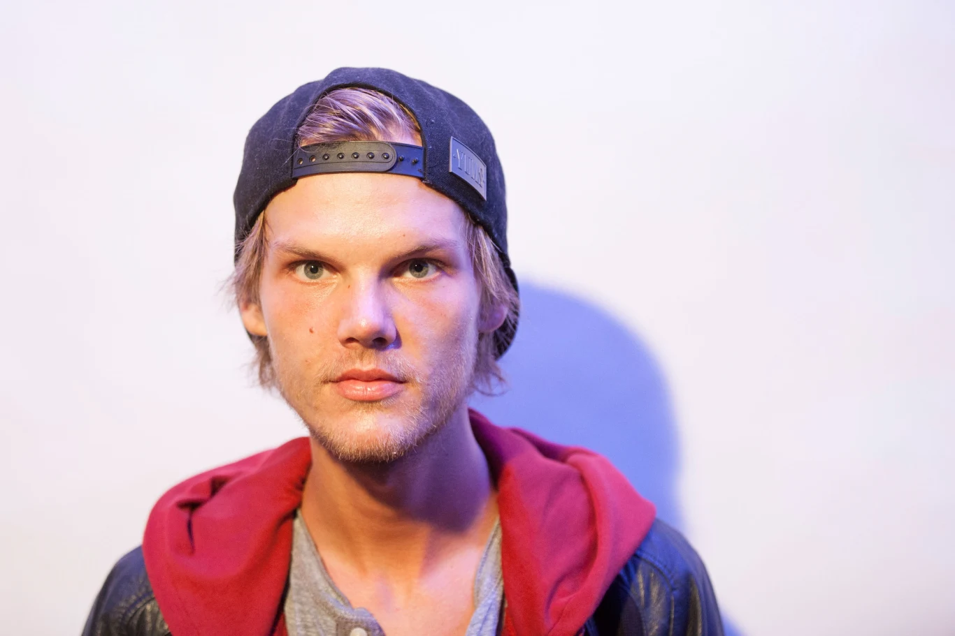 Avicii był jednym z najpopularniejszych twórców muzyki elektronicznej Mężczyzna w czapce z daszkiem patrzy prosto przed siebie, ma jasne włosy. Tło jest jednolite i jasne, oświetlone tak, że tworzy cień na ścianie. Ubrany jest w bluzę z kapturem, kurtkę i szary t-shirt.