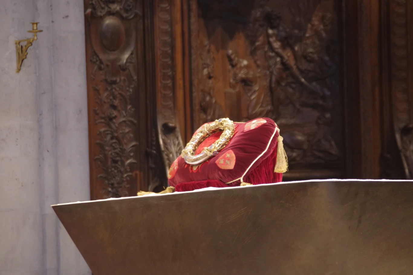 Poduszka z bogatymi zdobieniami w kolorze czerwonym, leżąca na drewnianym ołtarzu z rzeźbionymi panelami. Całość znajduje się w wnętrzu o ozdobnym i klasycznym charakterze.