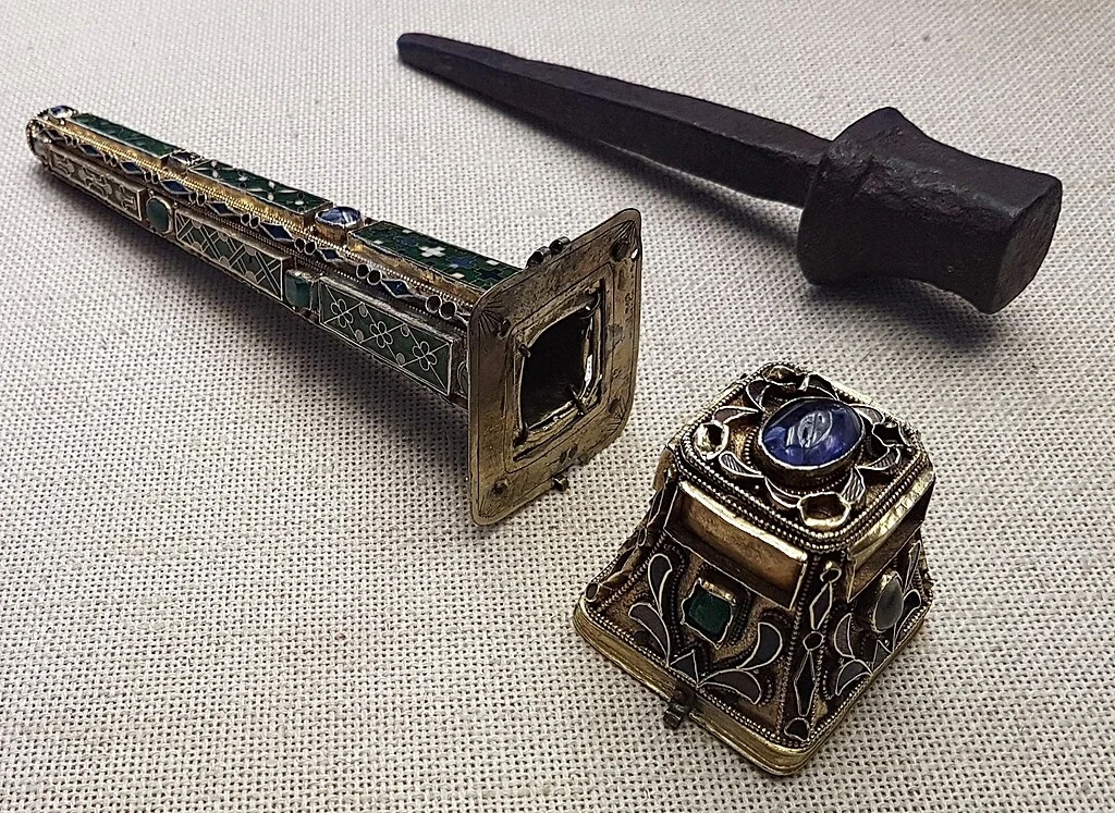 Zabytkowe przedmioty o ornamentacyjnych detalach, w tym bogato zdobiony nóż z rękojeścią pokrytą zielonymi emaliami oraz ozdobny przedmiot z metalu i kamieniami szlachetnymi. Na tle beżowego materiału.