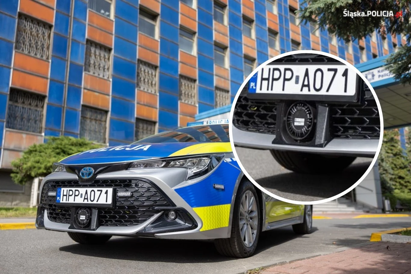 Na zderzakach radiowozów policyjnych zamontowano dziwny element. Co to jest? Radiowóz policyjny marki Toyota parkuje przed nowoczesnym budynkiem o niebiesko-pomarańczowej fasadzie, z wyraźnym zbliżeniem na tablicę rejestracyjną i umieszczoną pod nią kamerę monitorującą.