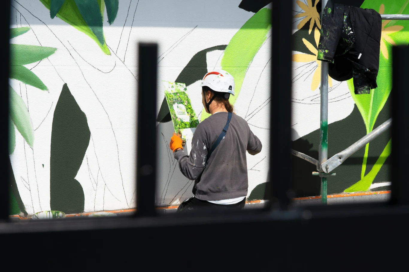 Osoba malująca mural w kasku ochronnym i rękawicach, stojąca na rusztowaniu. W tle widać abstrakcyjny wzór w roślinne motywy.