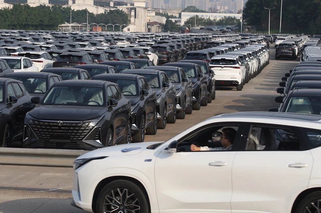 Rozległy parking pełen nowych samochodów, ustawionych w równych rzędach. Głównie ciemne pojazdy, z przodu biały SUV z kierowcą w środku kierujący się w lewo.