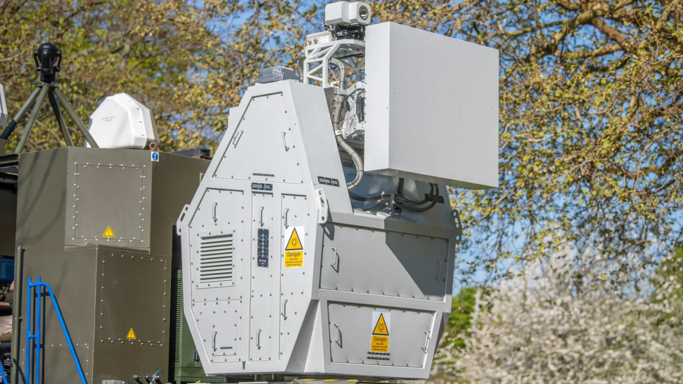Armia Wielkiej Brytanii przetestowała broń, która natychmiast może strącać masę dronów Zaawansowane urządzenie technologiczne zamontowane na zewnątrz. Wielkogabarytowa konstrukcja z panelami i wskaźnikami ostrzegającymi. Obok urządzenia widać fragment otaczających drzew.